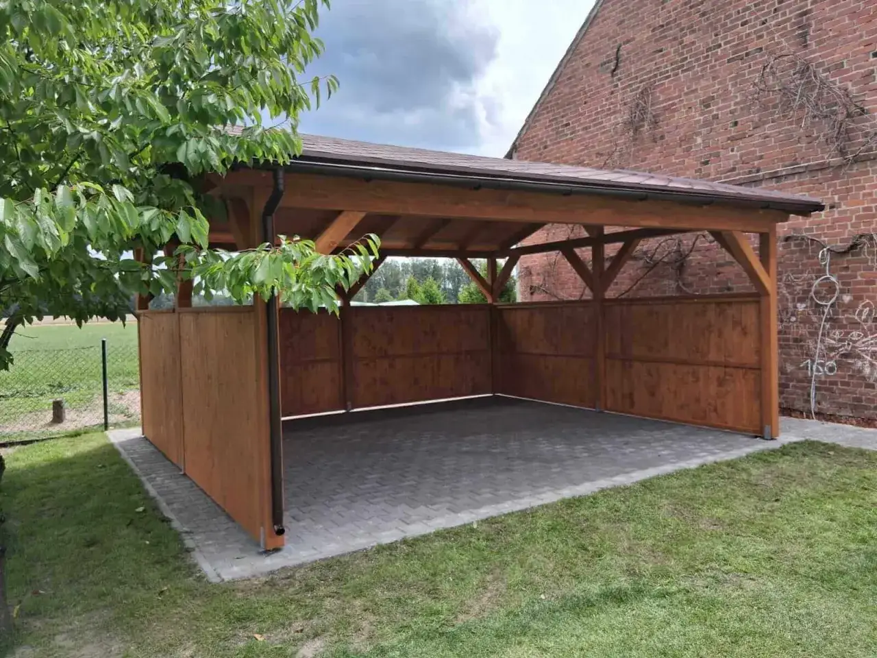 Drewniana wiata w granicy działki, zadaszona i częściowo osłonięta, stanowi schronienie dla pojazdu.