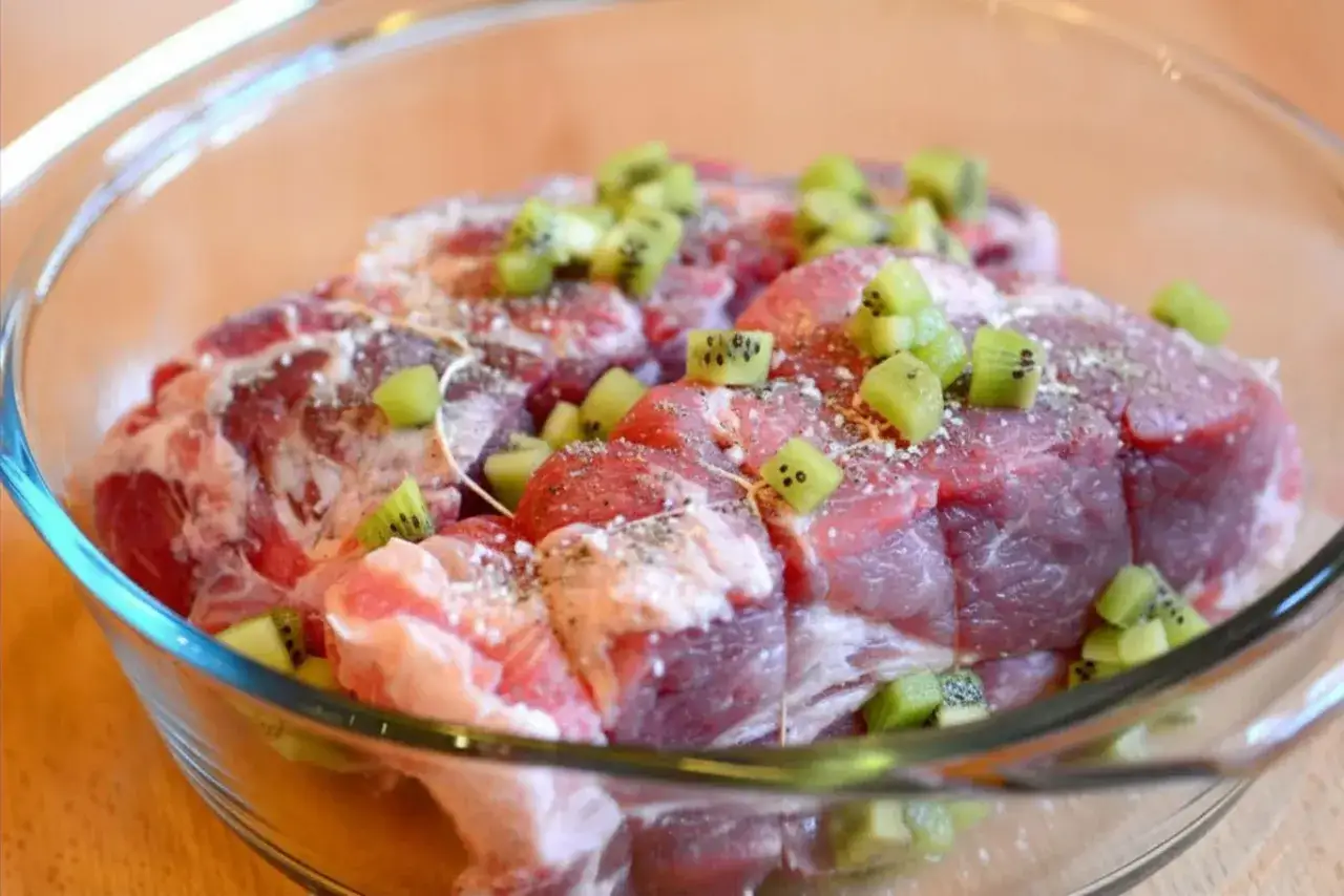 Surowe mięso wołowe w szklanej misce, posypane pieprzem i kawałkami kiwi. To naturalna marynata do wołowiny z kiwi zmiękczająca mięso.