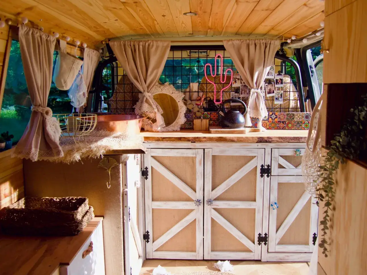 Przytulne wnętrze kampera z drewnianym sufitem, zasłonami i szafkami w stylu rustykalnym. Neonowy kaktus dodaje uroku.