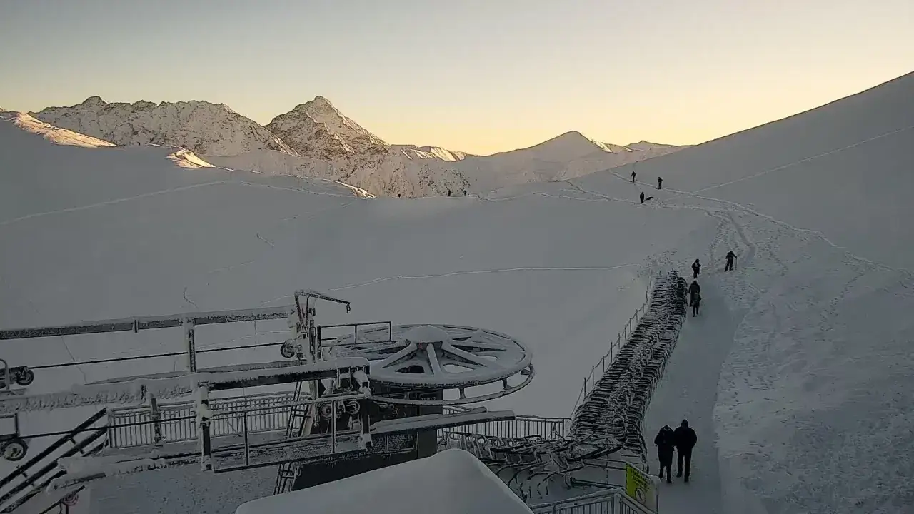 Widok z kamery na Kasprowy Wierch w Zakopanem. Śnieżne szczyty oświetlone słońcem, narciarze na stoku, kolejka linowa.