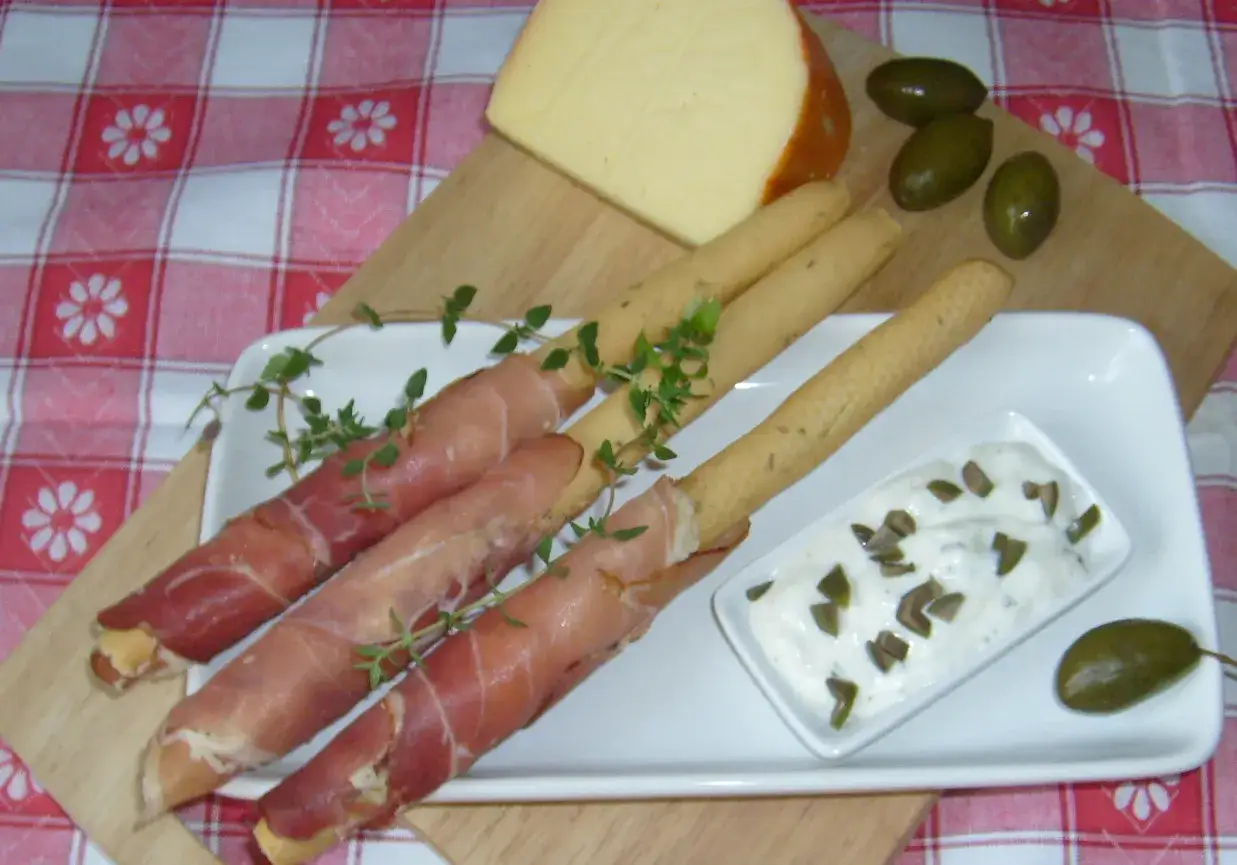 Grissini z szynką parmeńską, ser, oliwki i dip z kaparami na talerzu.