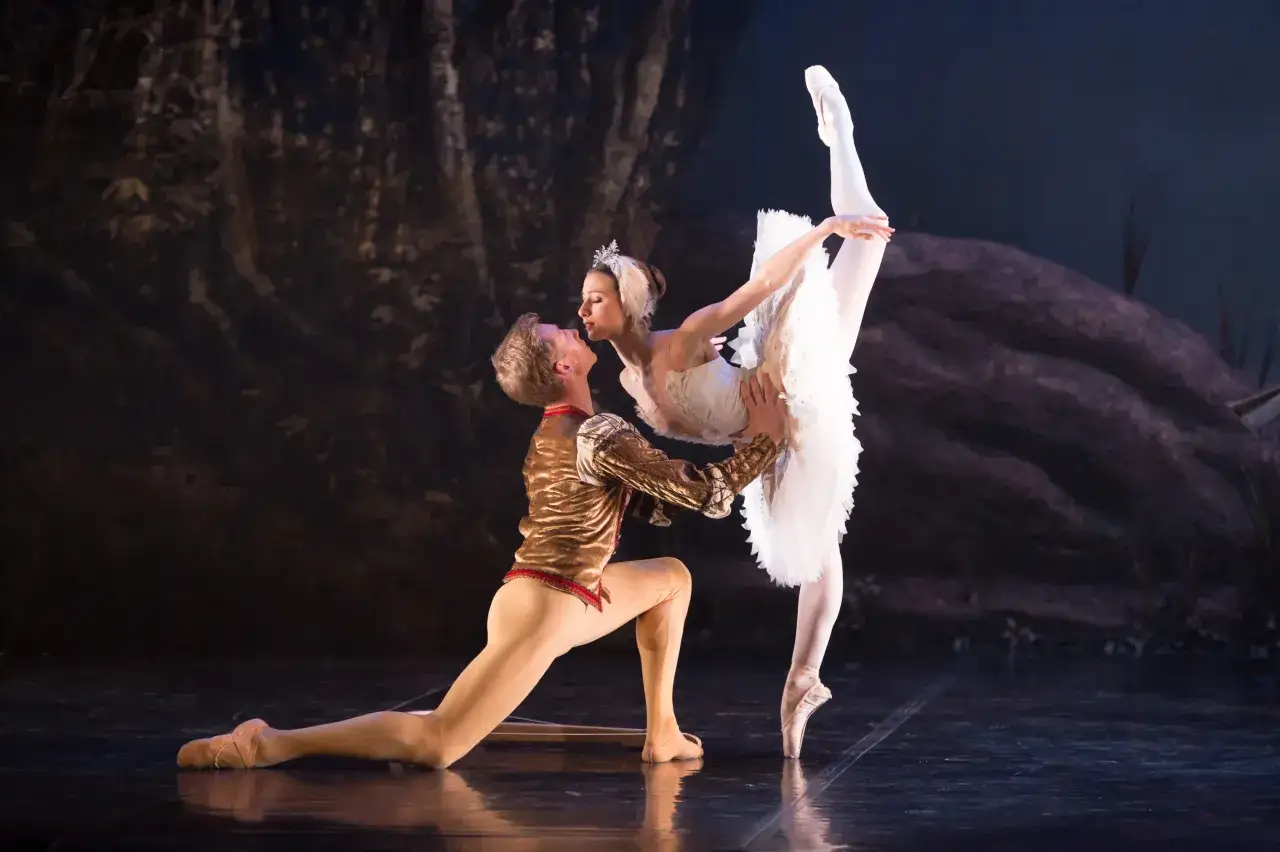 Baletnica w białej tutu i książę w złotym stroju wykonują skomplikowane pozycje baletowe na scenie.