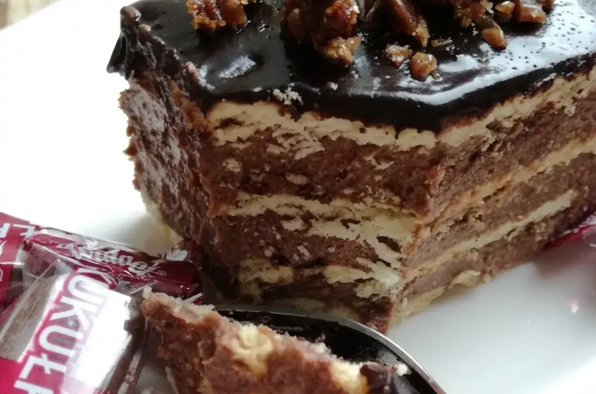 Czekoladowe ciasto kukułka z kremem i wafelkami, polane lukrem, z okruszkami na wierzchu. Obok opakowanie cukierków Kukułka.