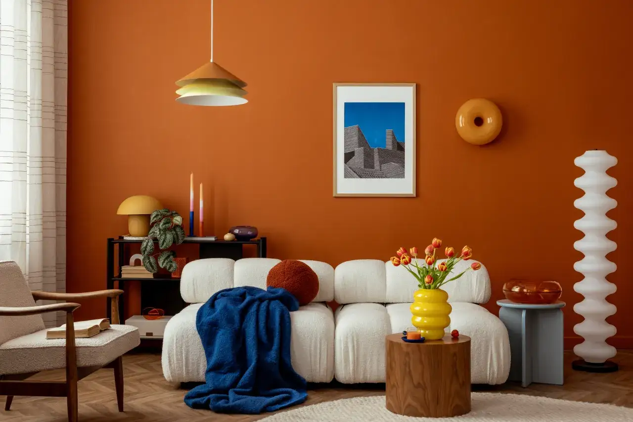 Inspiracja, jak pomalować ściany w salonie: ciepła pomarańcza, biała kanapa, drewniane meble i kolorowe dodatki tworzą przytulną atmosferę.