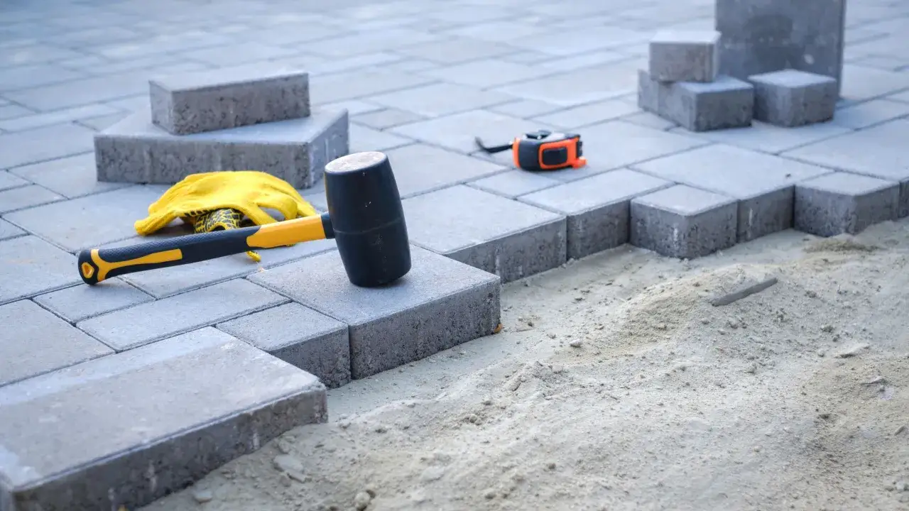 Młotek gumowy, rękawice i miarka leżą obok kostki brukowej. Przygotowanie podłoża z suchego betonu pod kostkę wymaga odpowiednich proporcji.