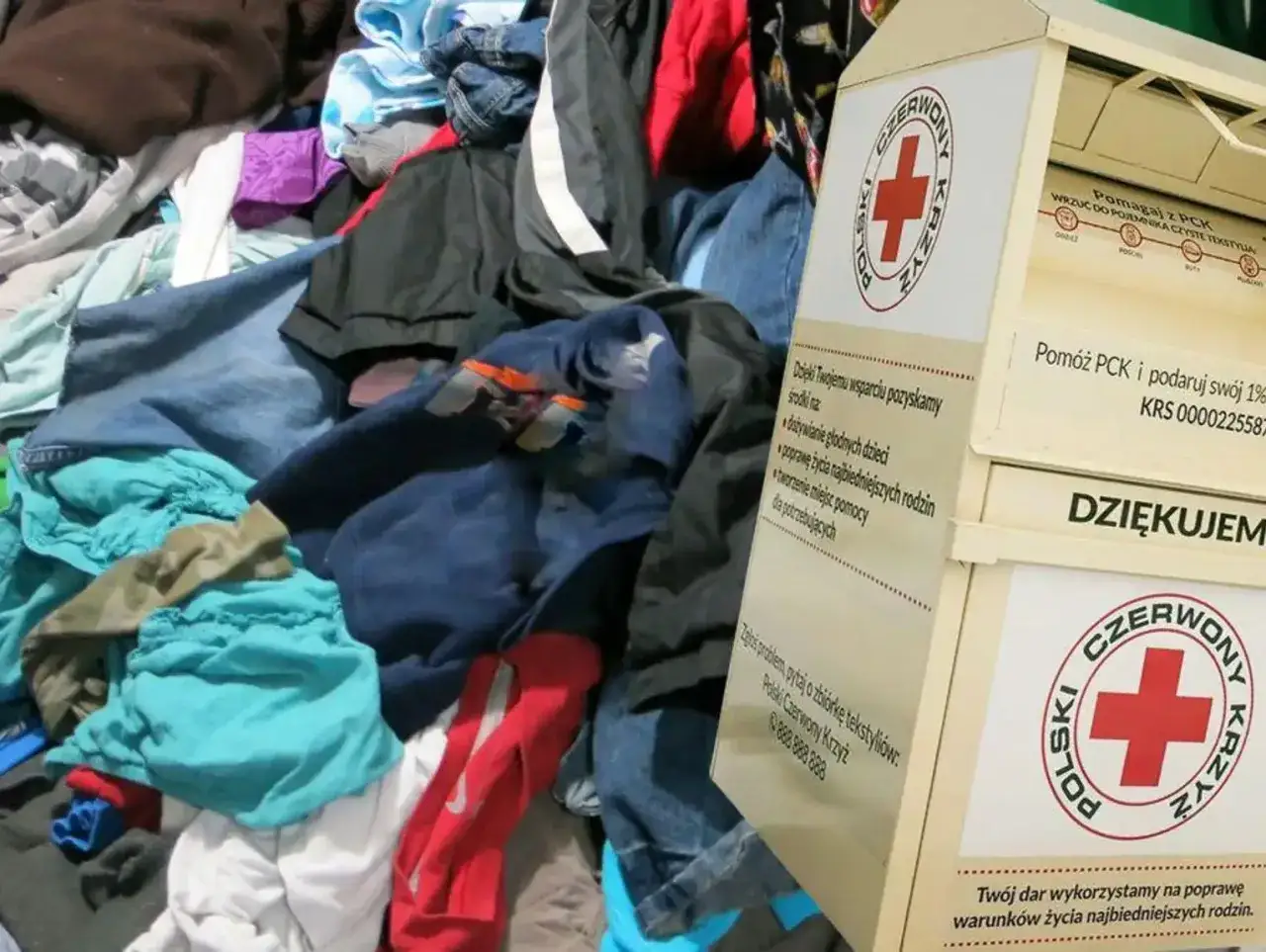 Pojemnik Polskiego Czerwonego Krzyża obok sterty ubrań. Tutaj można oddać ubrania dla potrzebujących, wspierając potrzebujących.