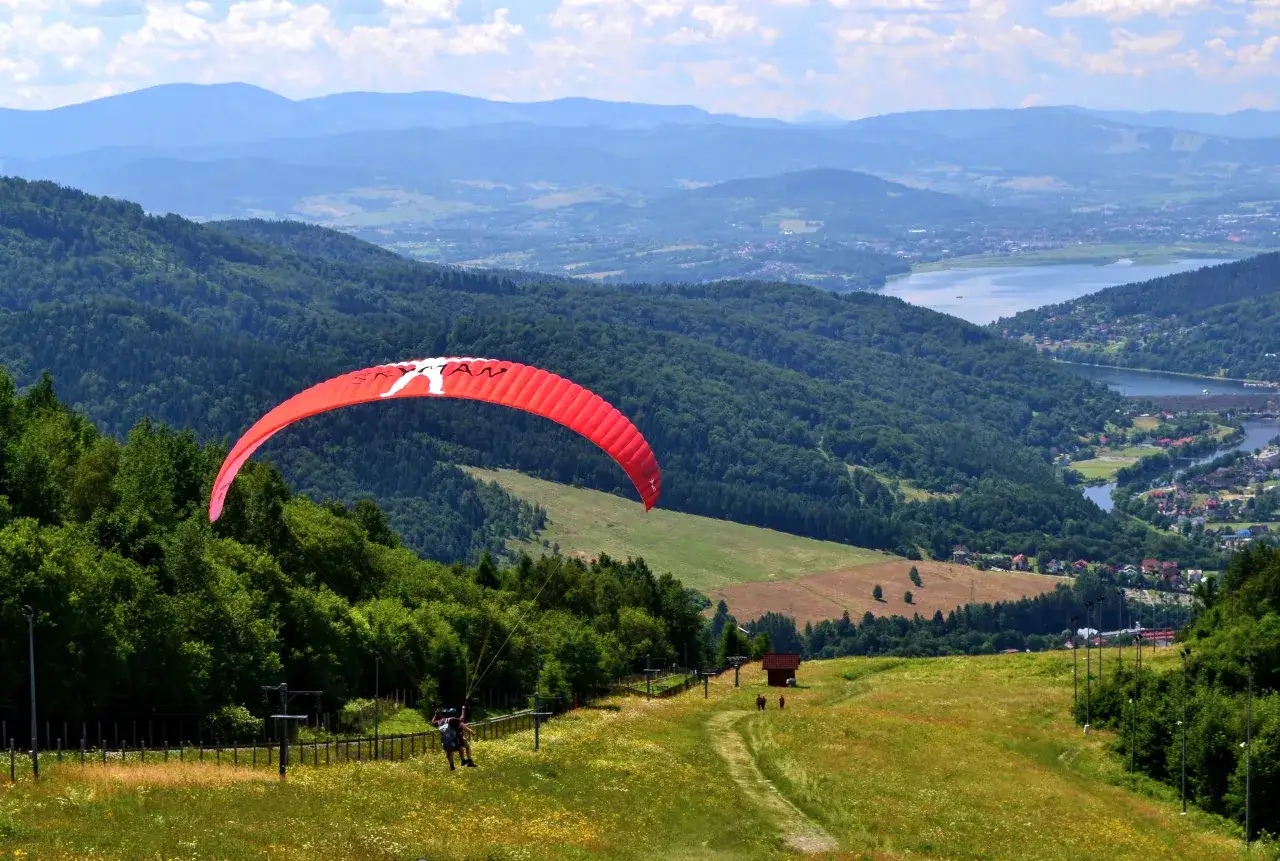 Paralotniarz szybuje nad zielonymi wzgórzami, gdzie jest góra Żar. W oddali jezioro i miasteczko.