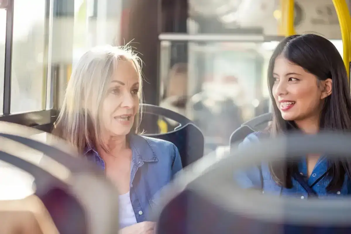 Dwie kobiety, starsza i młodsza, siedzą obok siebie w autobusie. Starsza kobieta z siwymi włosami opowiada coś młodszej, kt&oacute;ra uśmiecha się, słuchając. To miła rozmowa z nieznajomym.