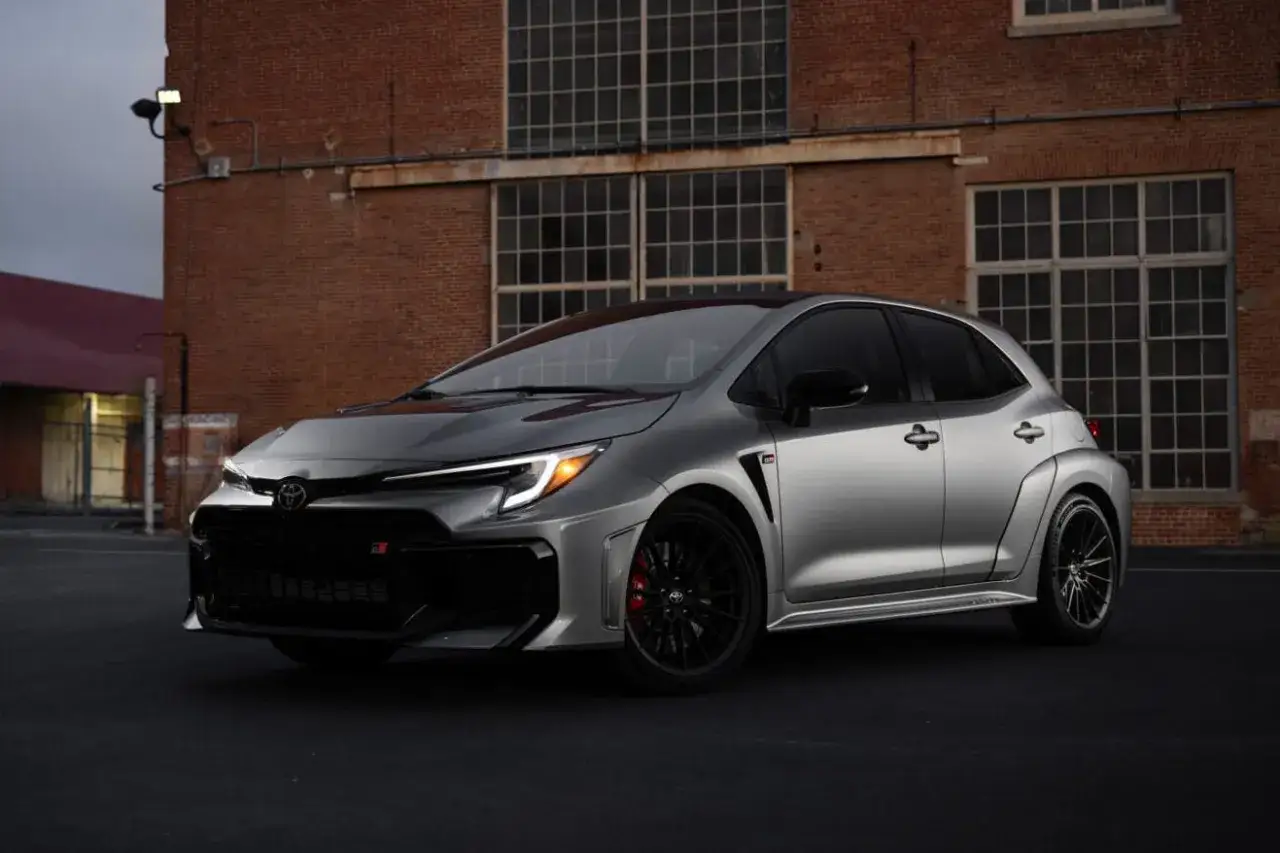 Srebrna Toyota Corolla GR, sportowy hatchback, zaparkowana przed ceglanym budynkiem z dużymi oknami.