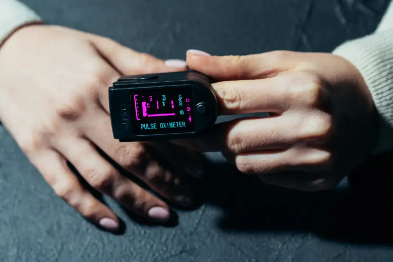 Dłoń z pulsoksymetrem pokazującym saturację 90%. Urządzenie mierzy poziom tlenu we krwi i tętno.