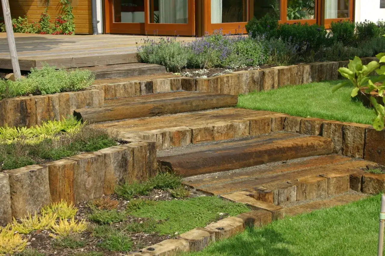 Drewniane schody prowadzą na taras na skarpie, otoczone zielenią i kwiatami.