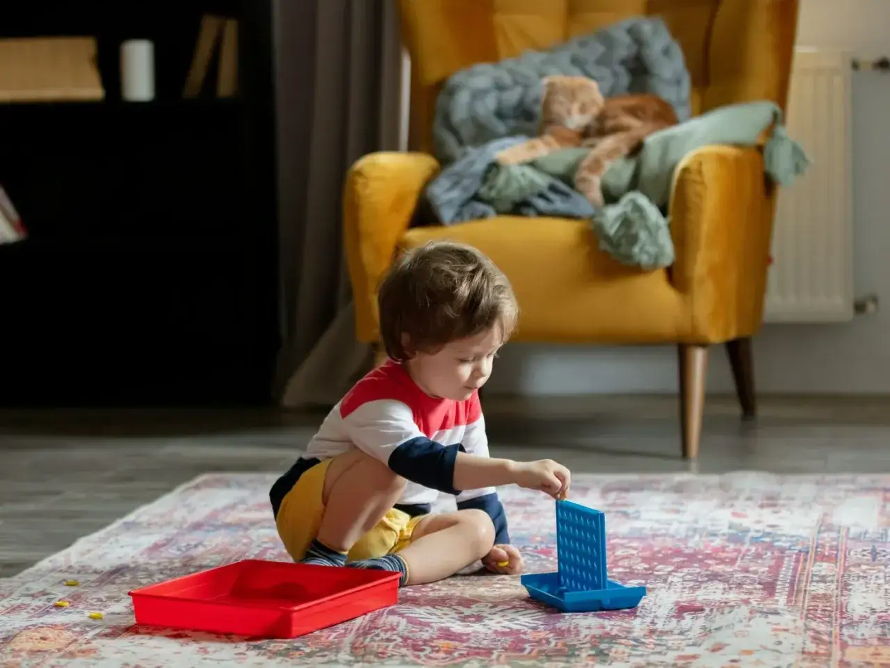 Chłopiec bawi się grą Connect 4 na dywanie. To idealne zabawy z 3 latkiem w domu, gdy koty odpoczywają na kanapie.