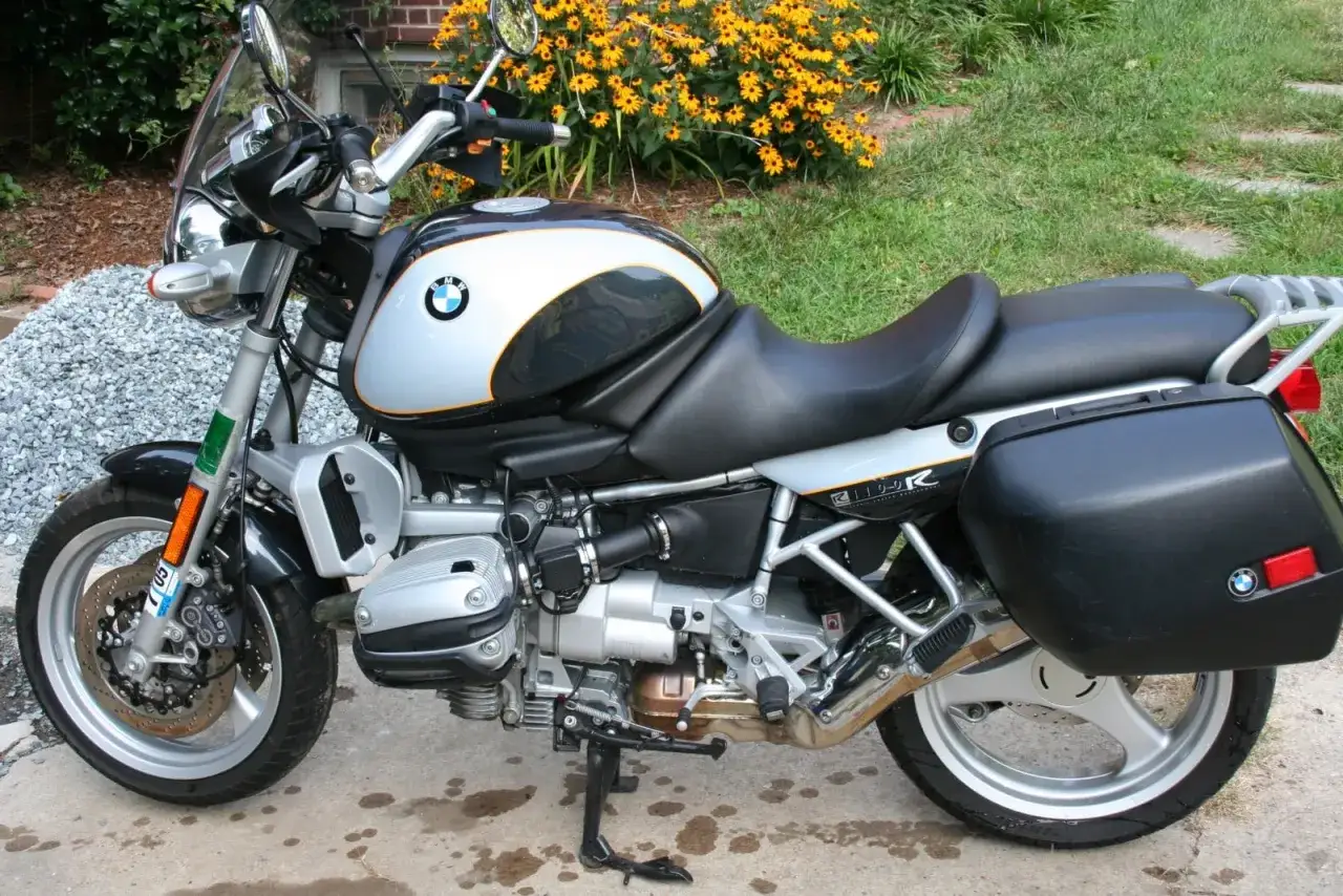Srebrno-czarny motocykl BMW R 1100 R z kuframi, zaparkowany na podjeździe, w tle ż&oacute;łte kwiaty.