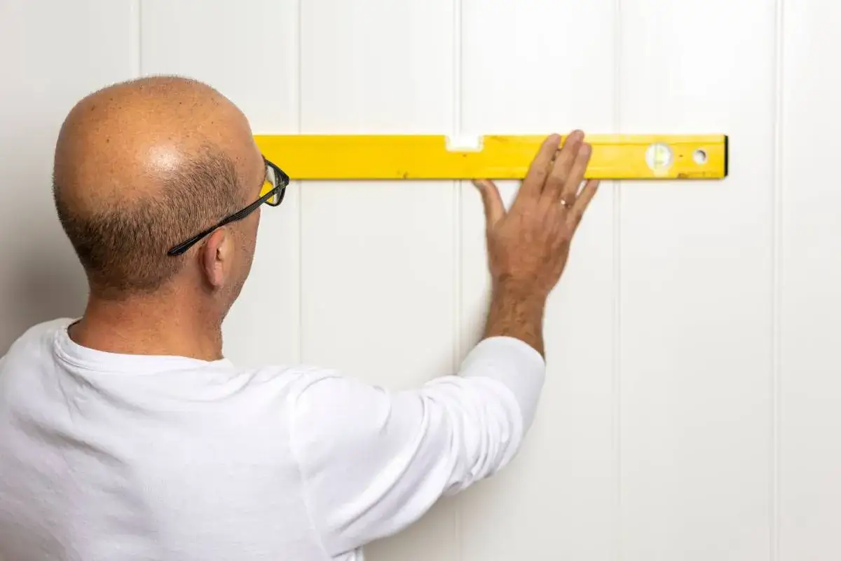 Mężczyzna z poziomicą sprawdza r&oacute;wność ściany. Planuje montaż paneli PCV, zastanawiając się, czym ciąć panele pcv, by uzyskać idealne dopasowanie.