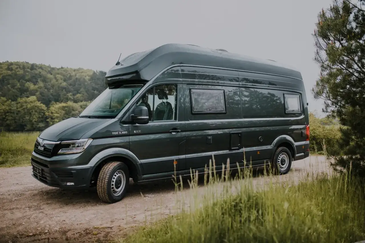 Ciemnozielony van camper MAN TGE 3.180 4x4 zaparkowany na leśnej drodze, gotowy na przygodę.