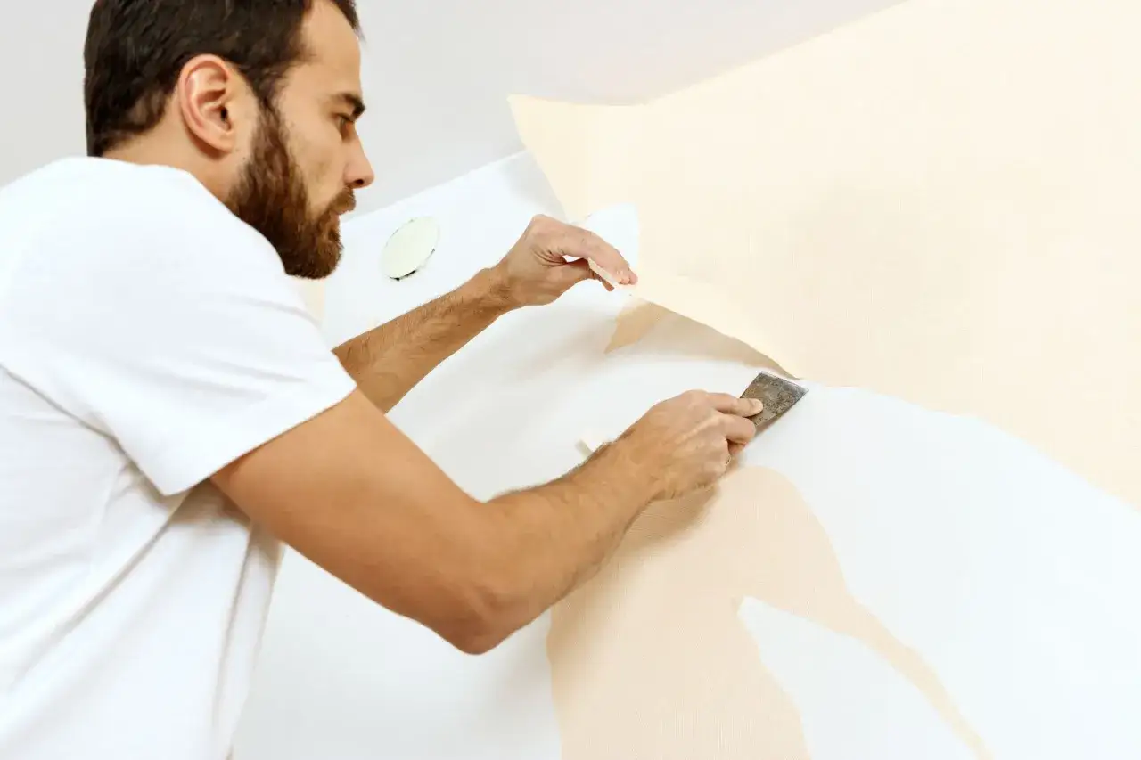 Mężczyzna z brodą, w białej koszulce, używa szpachelki, by pokazać, jak zerwać tapetę.