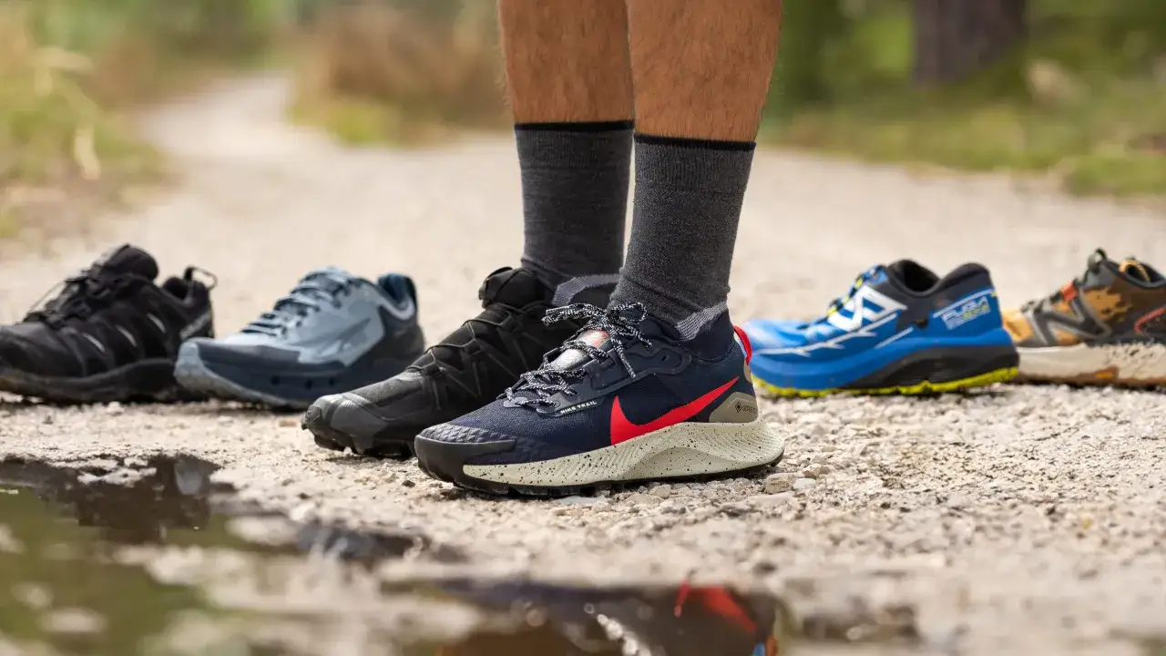 Buty trailowe co to? R&oacute;żne modele but&oacute;w do biegania w terenie, od czarnych po niebieskie, stoją na kamienistej ścieżce.