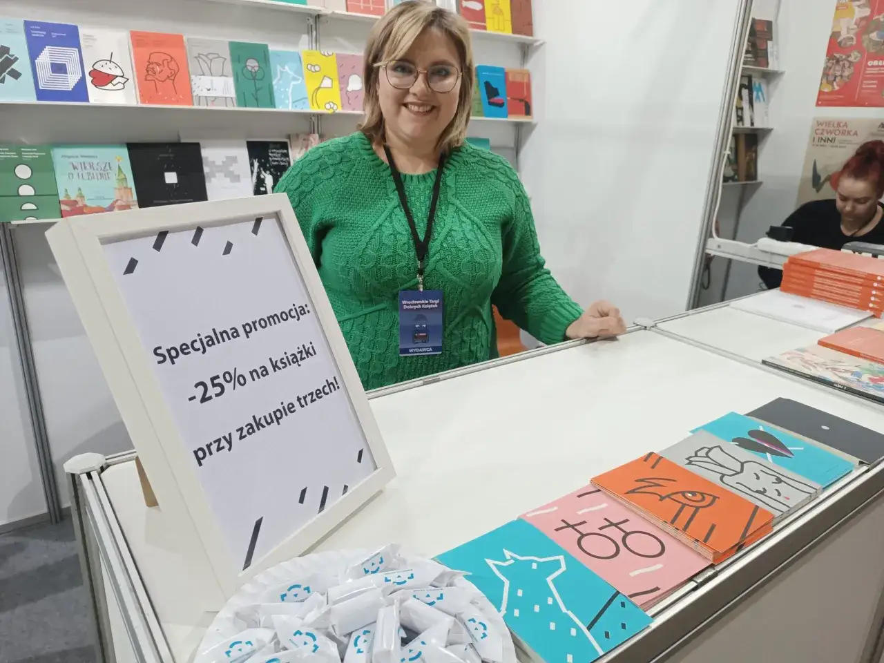 Kobieta w zielonym swetrze na stoisku z książkami, promująca wydawnictwa w Polsce. Promocja -25% na książki przy zakupie trzech.