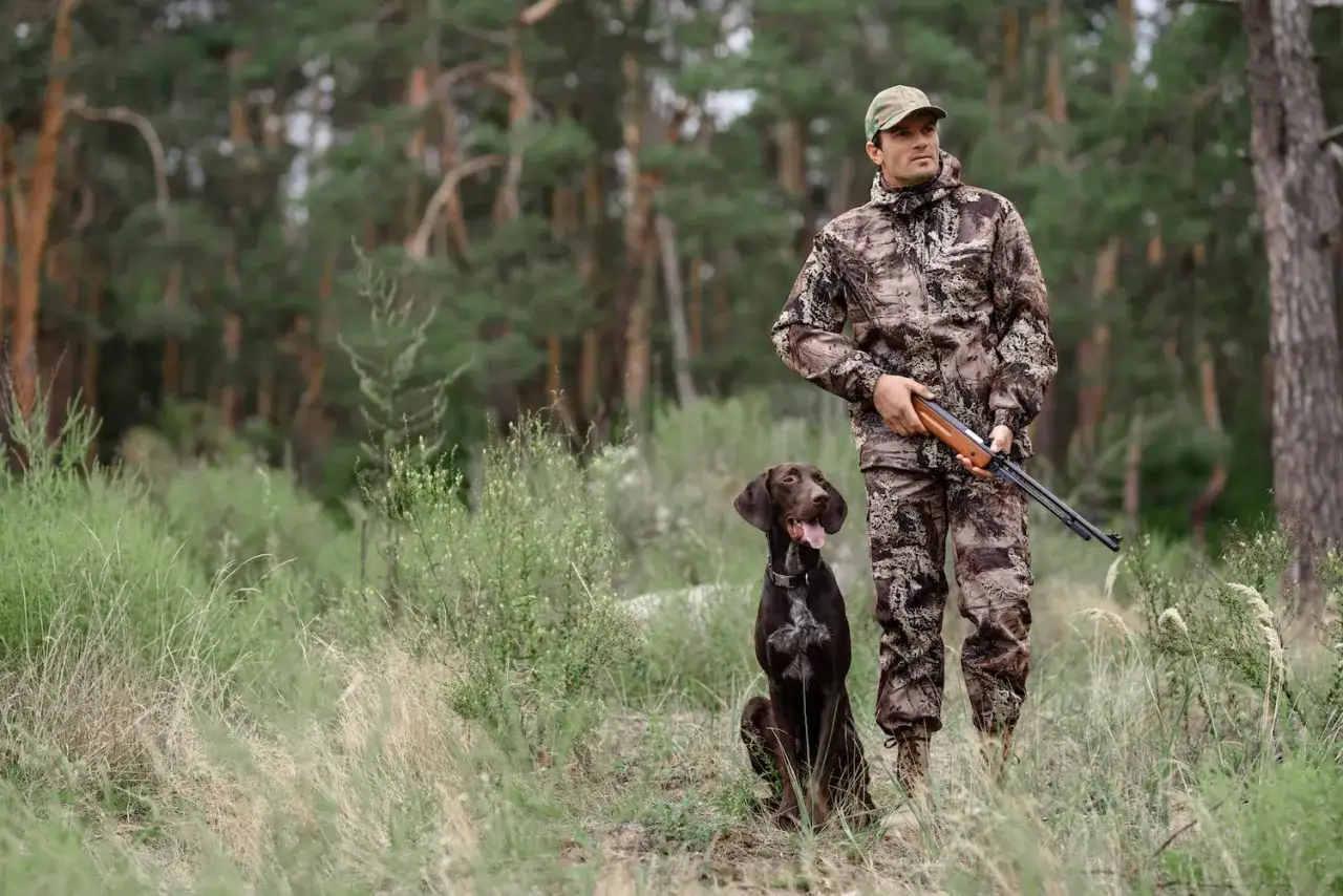 Myśliwy w kamuflażu z psem i strzelbą w lesie. Czy myśliwy może sam polować? Tak, z wiernym towarzyszem.