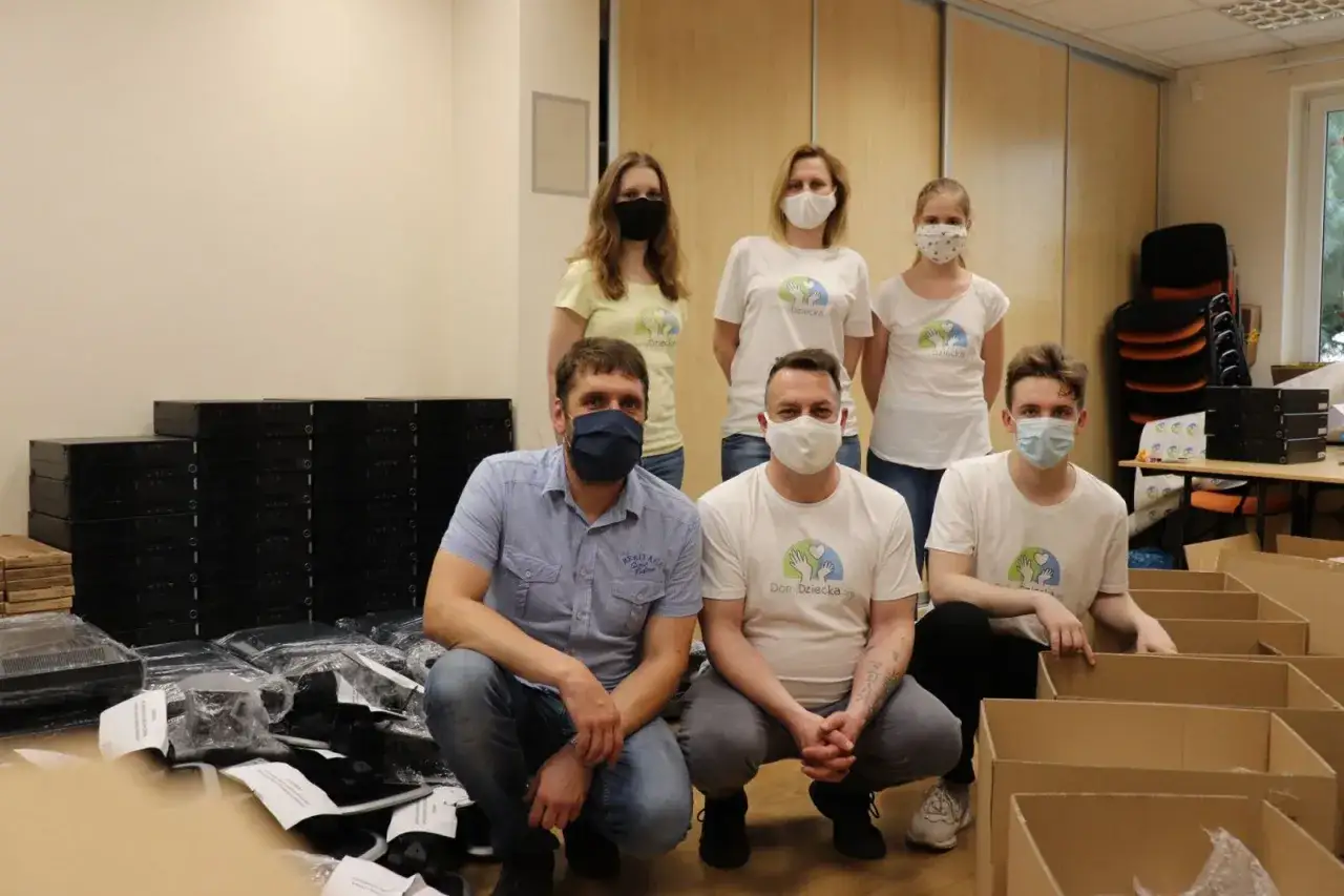 Grupa osób w maseczkach przygotowuje paczki. Domy dziecka przyjmują ubrania, co widać po stosach odzieży i kartonach.