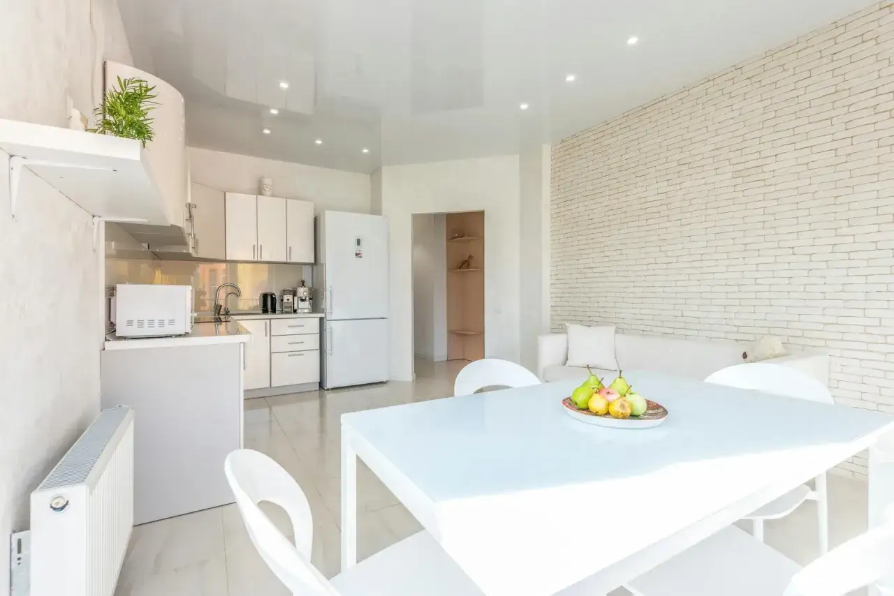 Jasna kuchnia z białym stołem i krzesłami. Na ścianie przy stole i krzesłach w kuchni widać białą cegłę i kanapę.