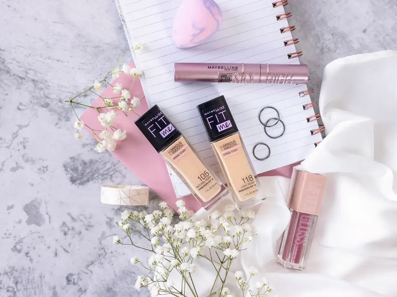 Maybelline Fit Me podkład rozświetlający, tusz do rzęs Sky High i błyszczyk Lifter Gloss na tle notatnika i kwiatów.