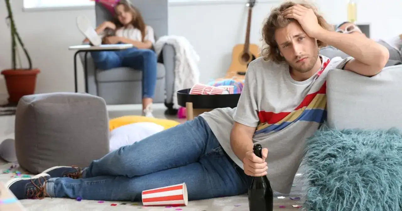 Młody mężczyzna z butelką w ręku, wyraźnie cierpiący na kaca, leży na podłodze wśr&oacute;d resztek imprezy. Obok niego kubek z napisem 