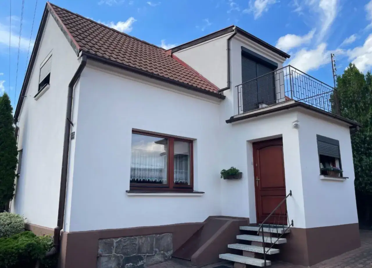 Dom po audycie energetycznym czyste powietrze. Biała elewacja, brązowy dach, drewniane okna i drzwi. Balkon z kutą balustradą.