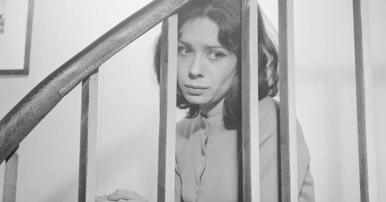 Lucyna Brusikiewicz spogląda zza drewnianych szczebelków schodów, jej wyraz twarzy pełen jest zadumy.