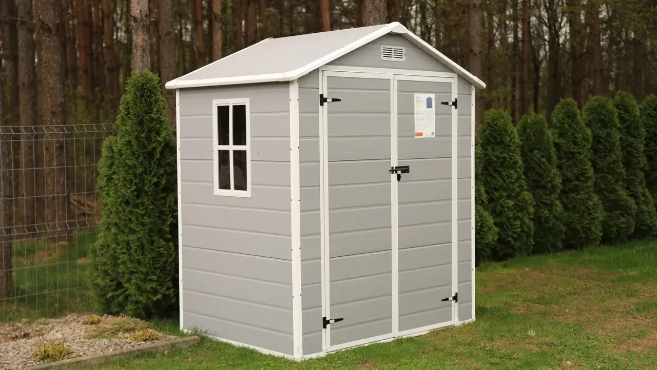 Szary domek narzędziowy z oknem i drzwiami, otoczony zielenią. Zastanawiasz się, czy domek narzędziowy trzeba zgłaszać?