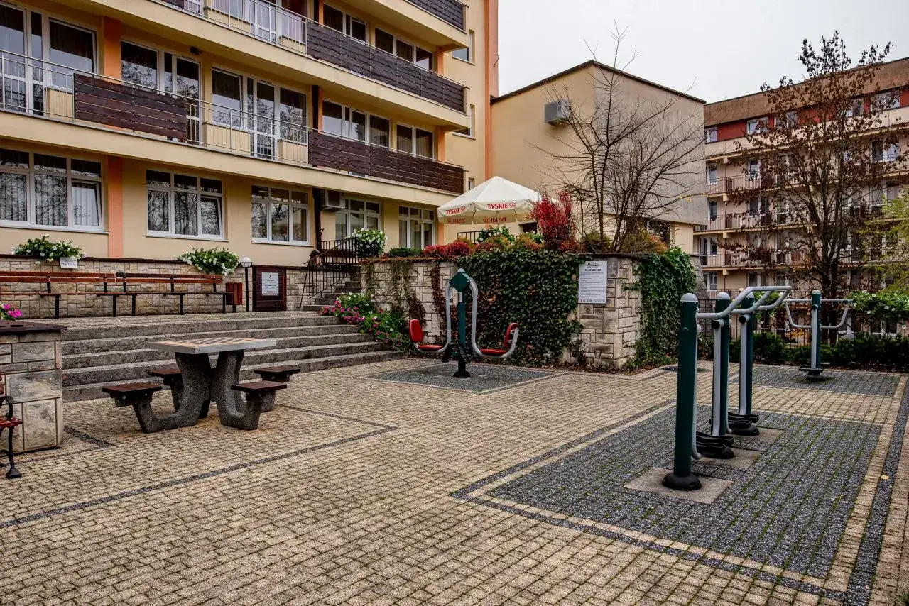 Sanatorium Leśnik Drzewiarz Krynica Zdr&oacute;j, ul. Podg&oacute;rna 1. Siłownia plenerowa i stolik do gry w szachy przed budynkiem.