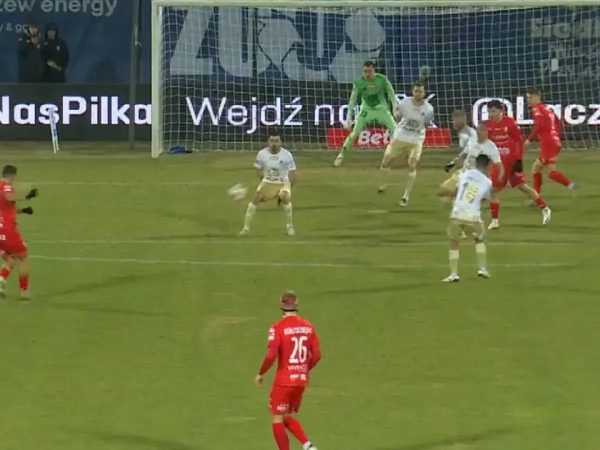 Piłkarze walczą o awans do Ekstraklasy. Bramkarz w zielonym stroju broni, a zawodnicy w białych i czerwonych koszulkach starają się zdobyć gola.