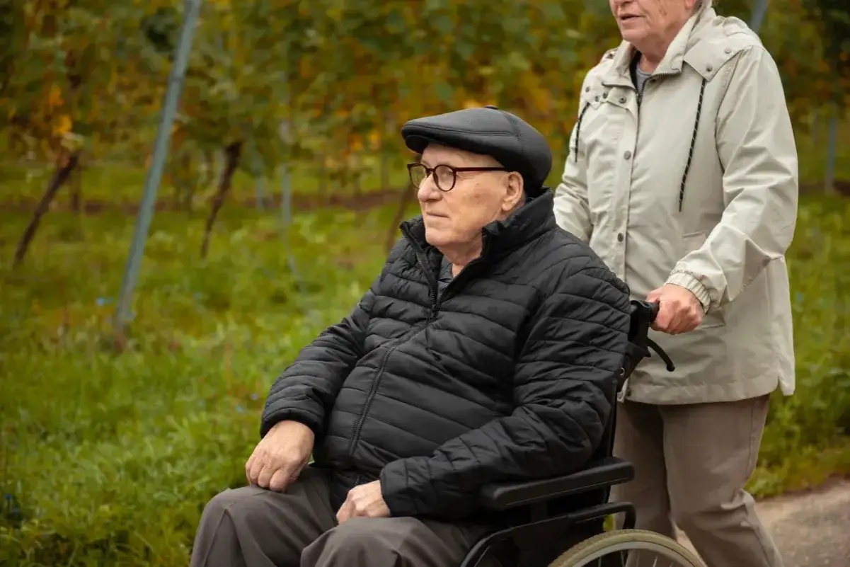 Starszy pan w wózku inwalidzkim, pchany przez kobietę. Informacje o tym, jak uzyskać status opiekuna osoby niepełnosprawnej, mogą pomóc w takich sytuacjach.