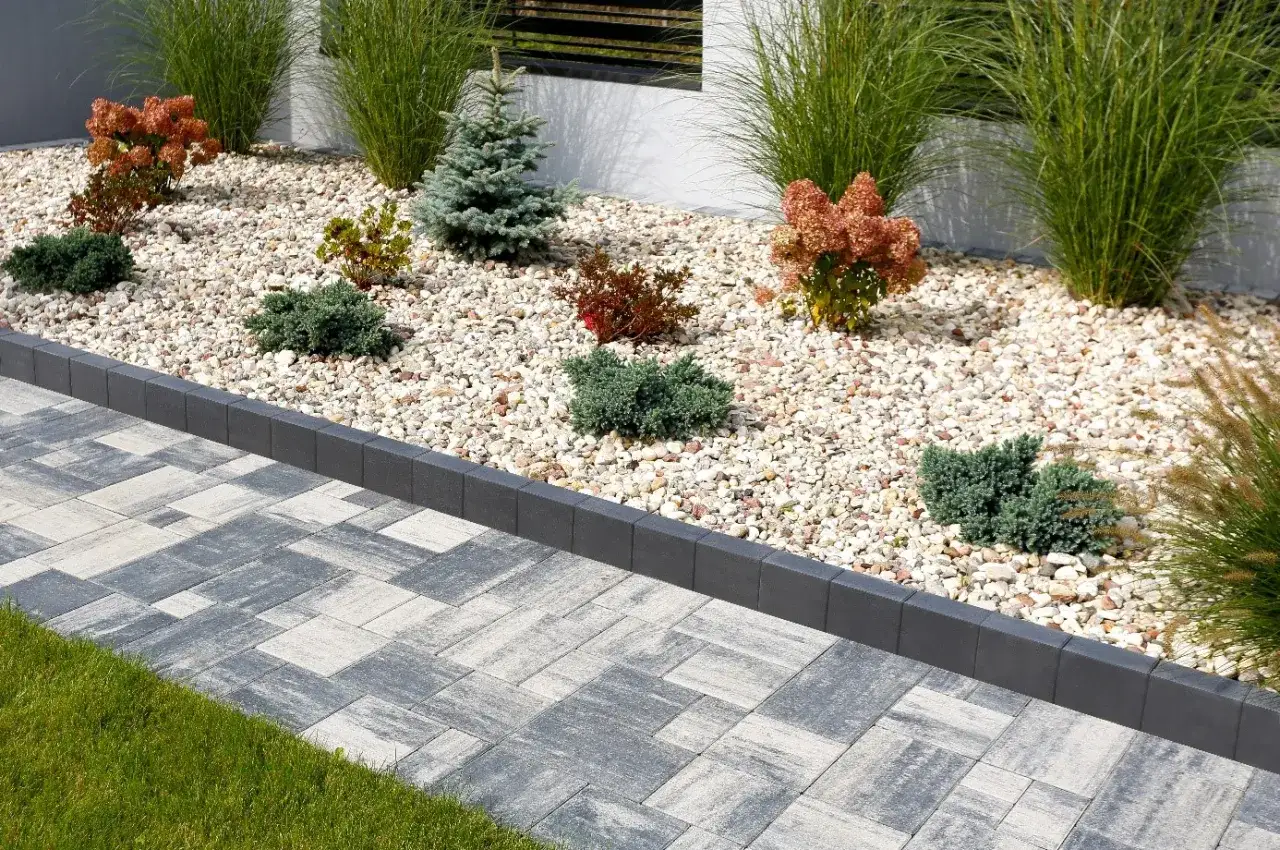 Kamienna ścieżka obok rabaty z iglakami i trawami ozdobnymi. Kamień jako materiał, którym ściółkować rabaty kwiatowe, tworzy nowoczesny wygląd.