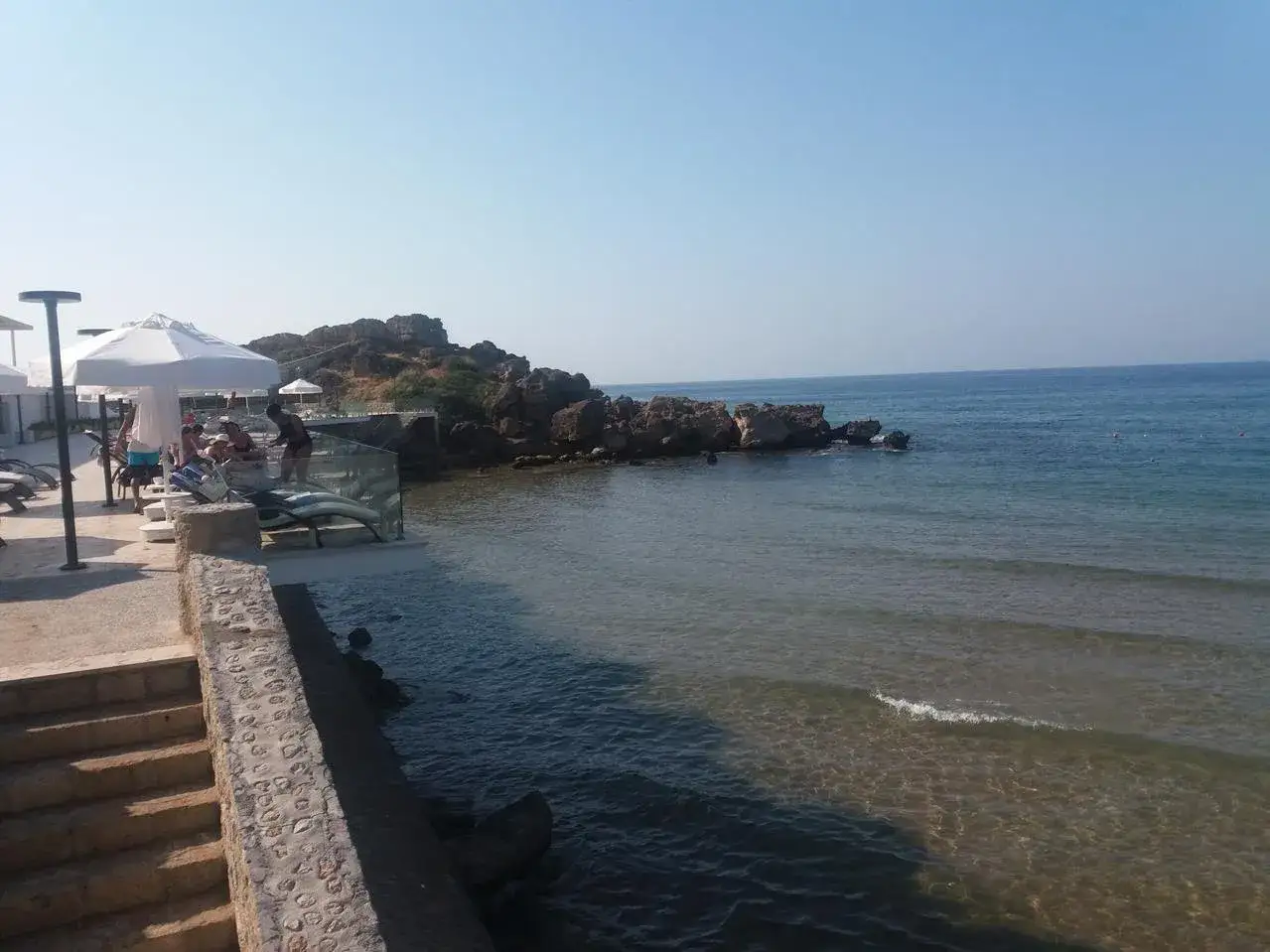 Relaks przy morzu w pobliżu **Św. Konstantyna i Eleny**. Widać tu kamieniste wybrzeże, turkusową wodę i leżaki pod białymi parasolami.