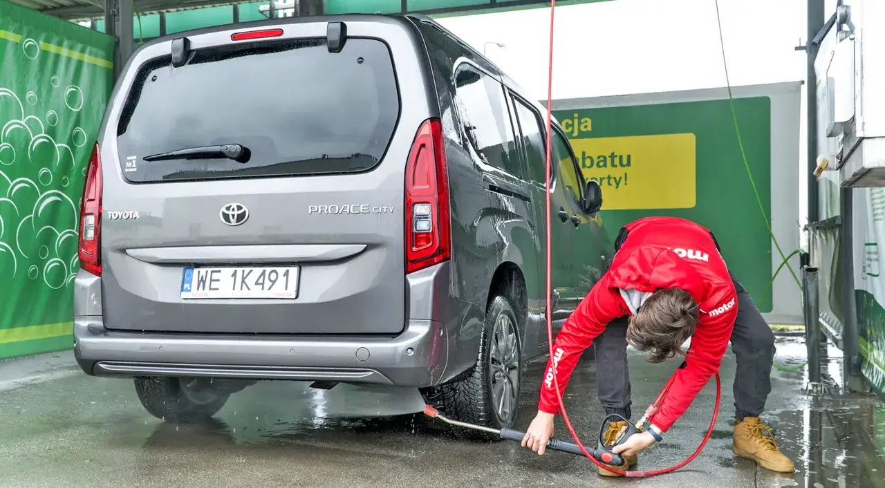 Chłopak w czerwonej kurtce pokazuje, jak myć auto na myjni bezdotykowej, spryskując Toyotę Proace City wodą pod ciśnieniem.