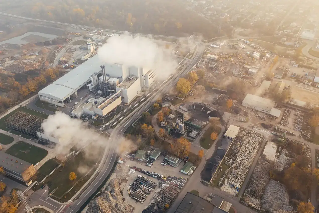 Widok z lotu ptaka na zakład przemysłowy z unoszącą się parą, kt&oacute;ra wpływa na jakość powietrza. Widać drogi, składy materiał&oacute;w i budynki.