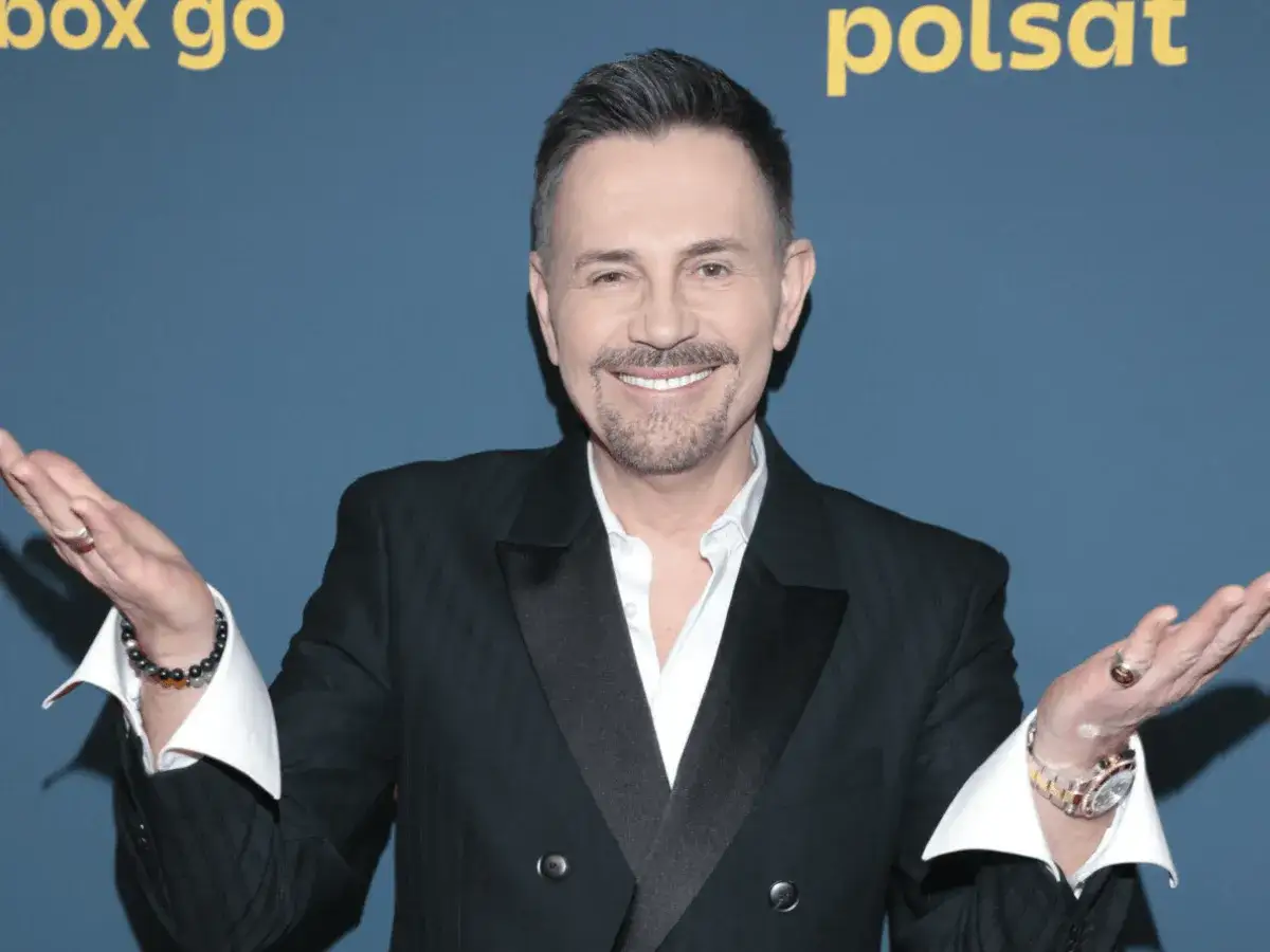 Uśmiechnięty Krzysztof Ibisz w eleganckim garniturze, z gestem otwartych dłoni. Jego styl i prezencja sugerują sukces, a wielu zastanawia się nad jego majątkiem.