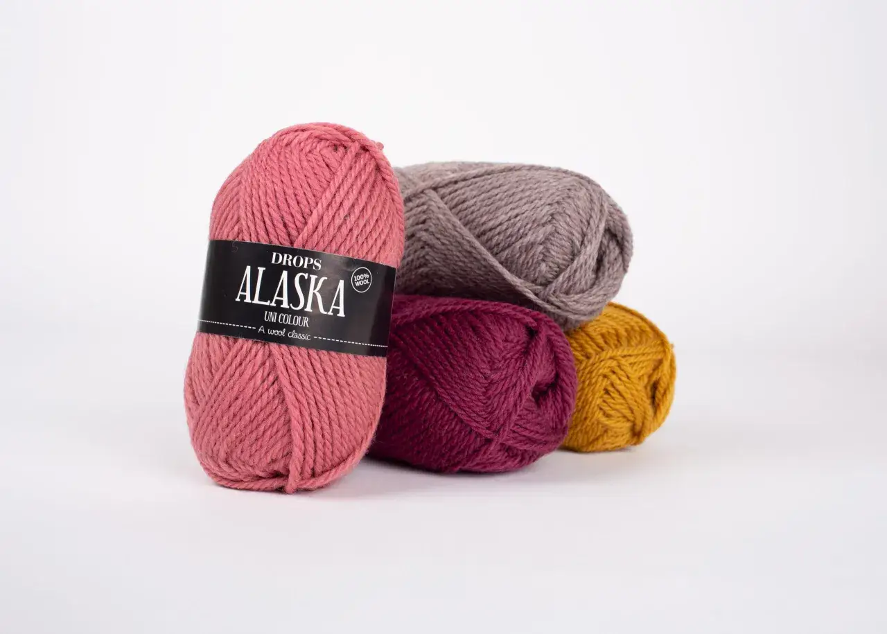 Kłębek różowej włóczki Drops Alaska z etykietą, obok inne kłębki w odcieniach szarości, bordo i musztardy. Idealne na czapkę i szalik.