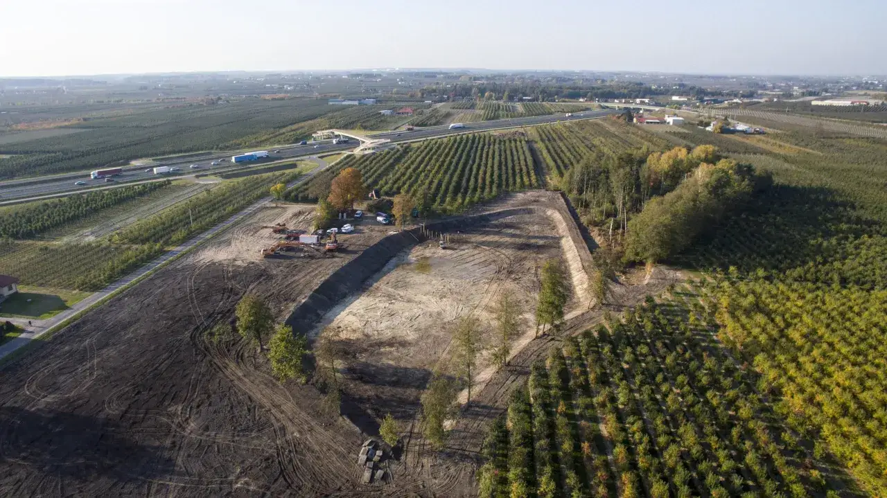 Budowa stawu w trakcie, koparki pracują. Zastanawiasz się, ile kosztuje wykopanie stawu? Obok autostrada i plantacje drzew.