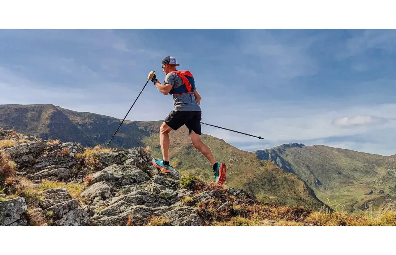 Mężczyzna biegnie po skalistym szlaku, używając kijk&oacute;w do biegania po g&oacute;rach. W tle rozciągają się zielone wzg&oacute;rza i błękitne niebo.