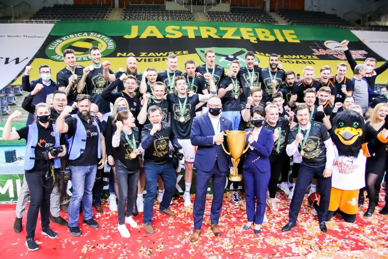 Drużyna siatkarzy świętuje zwycięstwo z pucharem. Na koszulkach widnieje napis 