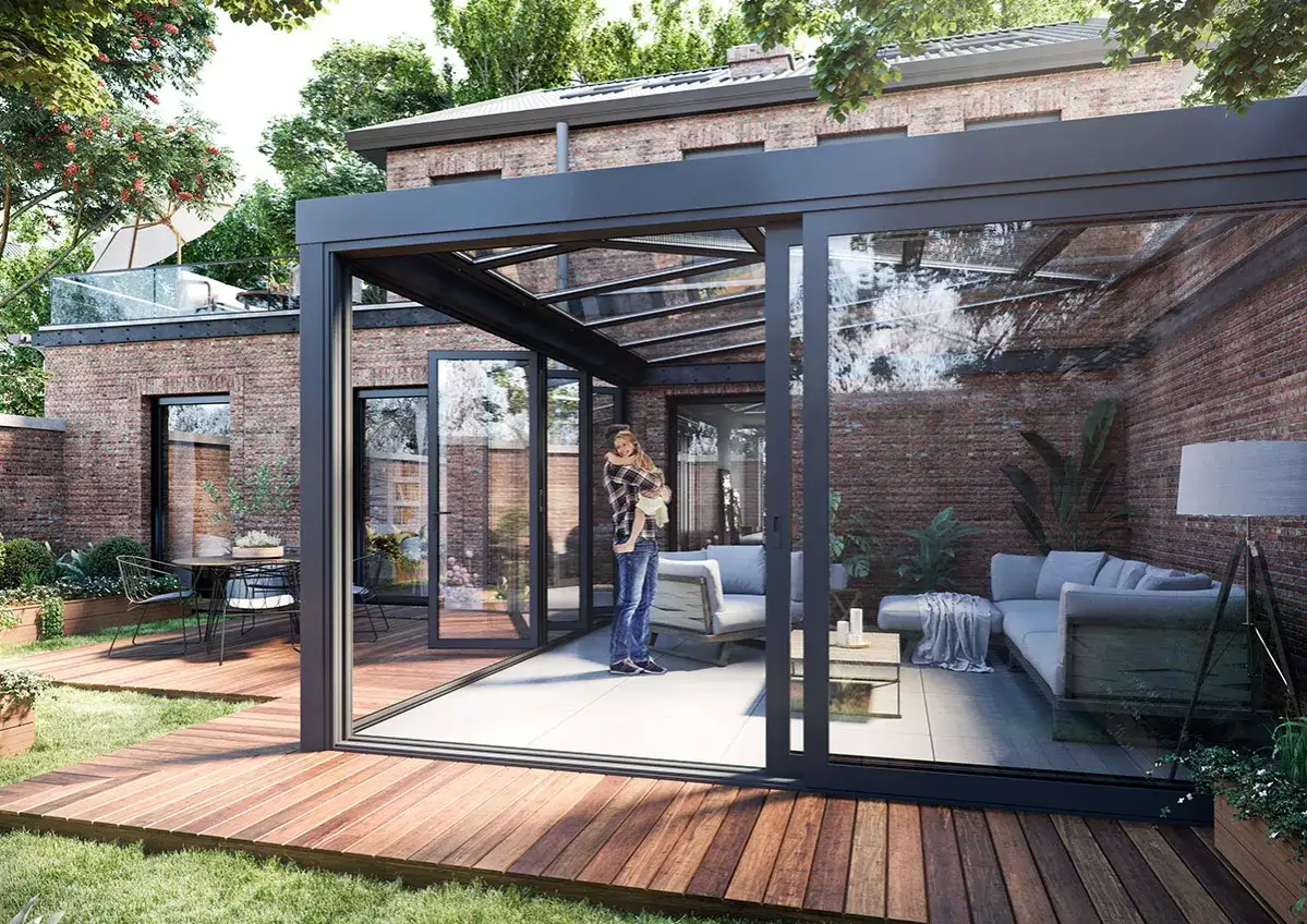 Nowoczesny wystr&oacute;j okna tarasowego z przeszklonym dachem i ścianami. Wewnątrz salon z sofami, stolikiem i lampą. Na zewnątrz drewniany taras i ogr&oacute;d.