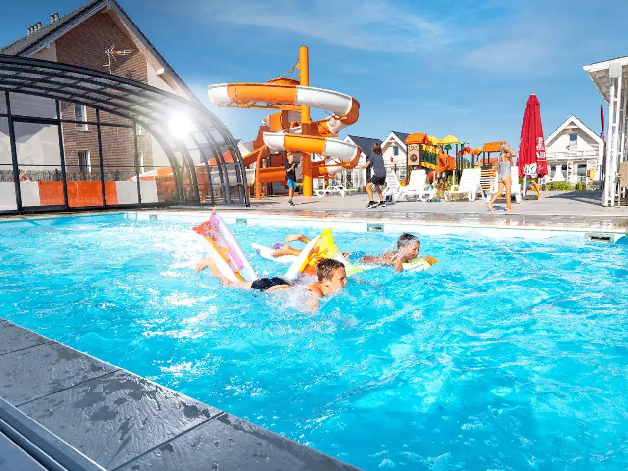 Dzieci pływają na dmuchanych zabawkach w basenie, w tle zjeżdżalnie wodne i domki. Idealne atrakcje dla dzieci nad morzem.