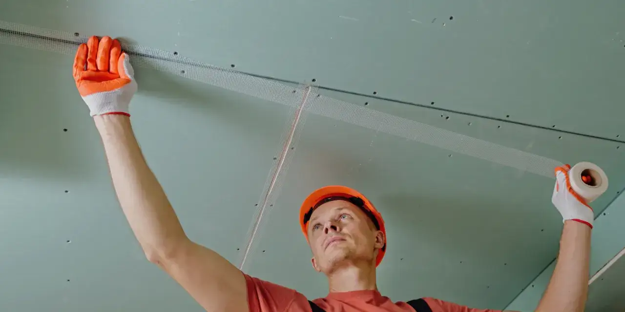 Budowlaniec w pomarańczowym kasku i rękawicach nakleja siatkę na połączenie płyt gipsowo-kartonowych. Czy można malować płytę GK bez gładzi? Tak, ale wymaga to odpowiedniego przygotowania.
