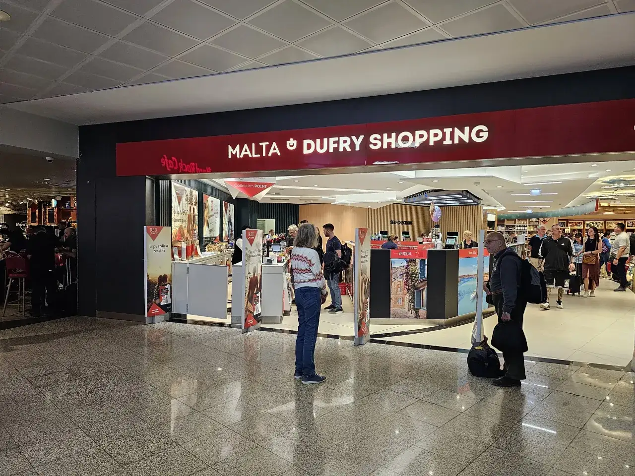 Sklep Malta Duty Free na lotnisku. Ludzie robią zakupy i czekają w kolejce do kasy samoobsługowej.