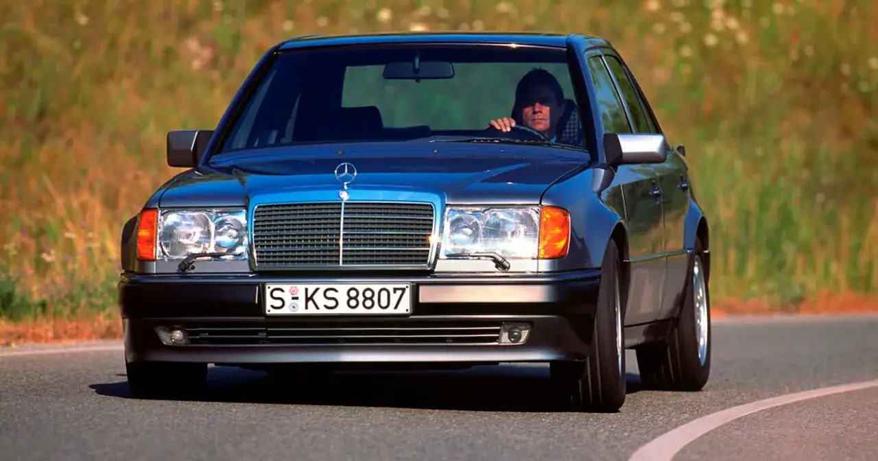 Klasyczny Mercedes W124 z kierowcą za kierownicą, jadący po krętej drodze wśród zieleni.