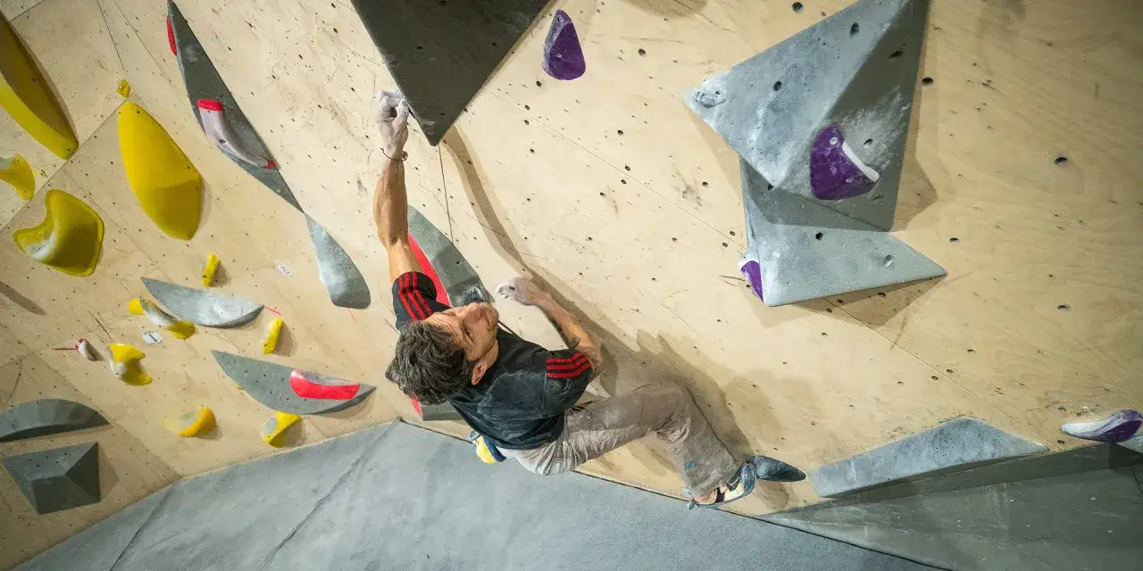 Mężczyzna w czarnej koszulce z czerwonymi pasami wspina się po drewnianej ścianie z kolorowymi chwytami. To jego pr&oacute;ba wspinaczki tradowej.