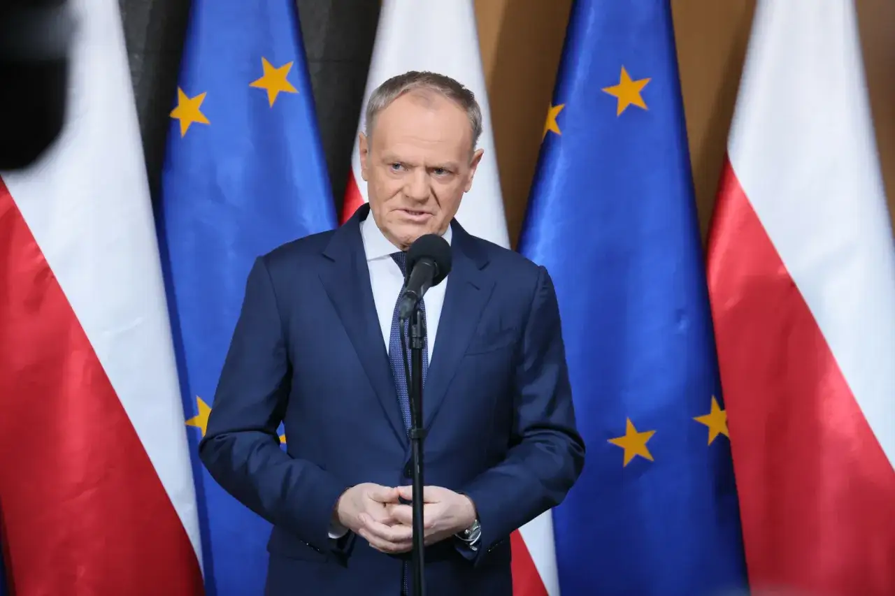Donald Tusk przemawia na tle flag Polski i UE. Sondaże nie dają pewności, czy rząd koalicyjny przetrwa 2026 rok w niezmienionym składzie.