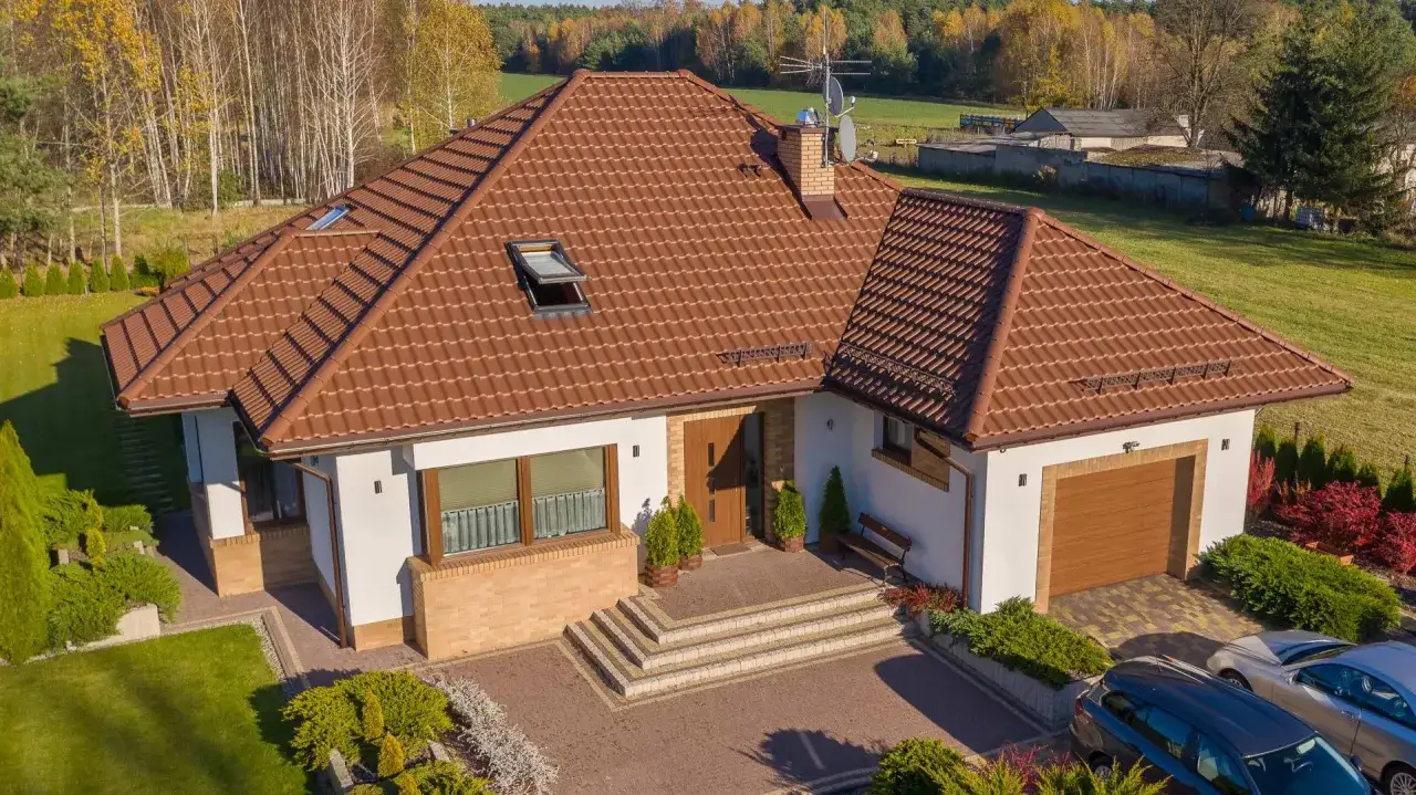 Dom z brązowym, ceramicznym dachem. Widać schody, podjazd, ogród i samochody. Ile dachówek na m2? Trudno ocenić, ale dach wygląda na solidnie pokryty.